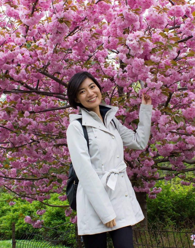 Fa Wang, a woman with short dark hair wearing a white coat, stands in front of a tree with pink blossoms.